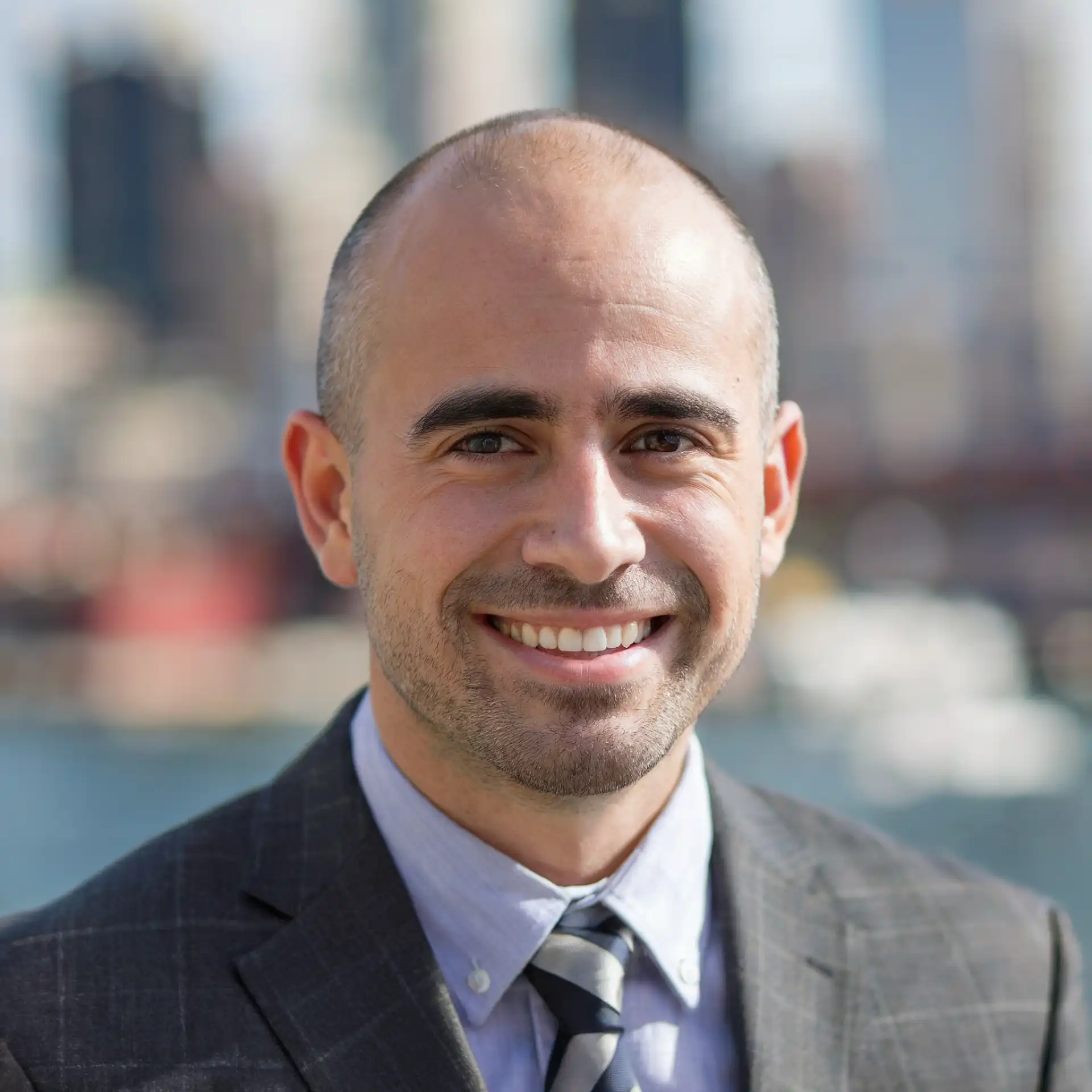 A man in a suit and tie smiles at the camera. The background shows a blurred cityscape with water and buildings.